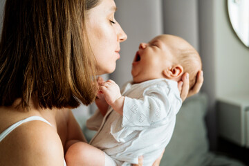A young mother is sitting on the bed and holding a baby in her arms.