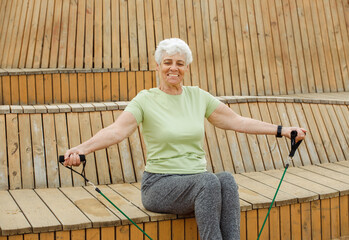 Old woman with short hair doing exercises outdoor , sitting on wooden bench using resistance rubber bands.