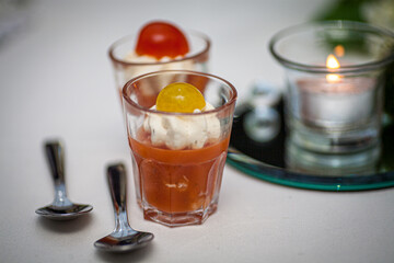 This image features an elegant appetizer presentation with a glass of creamy tomato soup topped with a dollop of rich cream and a cherry tomato garnish. The soup is served in a clear shot glass