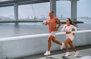 Focused and fit, a man and woman in trendy sportswear enjoy a seaside run, their athletic forms in harmony with the expansive bridge and tranquil waters framing their route.