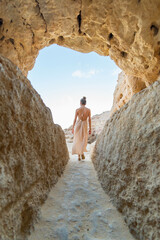 Journey Through the Natural Archway: A Low-Angle, Vertical Shot of a Woman in a Flowing Dress, Walking Away from the Camera Through a Sandstone Arch in a Desert Landscape