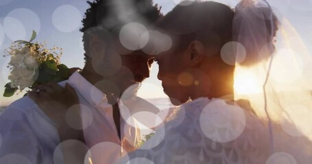 Animation of light spots over happy african american bride and groom embracing on beach at wedding