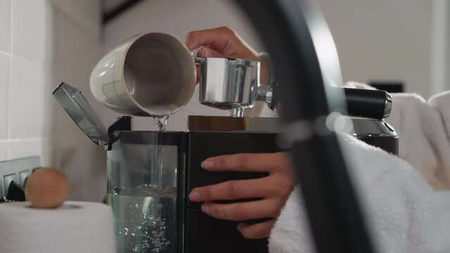 Woman pours water into coffeemaker tank in kitchen closeup. Lady fills water tank using cup preparing hot drink in morning. Servicing coffee machine