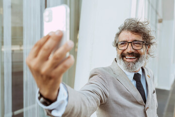 Smiling senior businessman, wears suit, eyeglasses, beard and curly gray hair, takes a selfie with mobile phone outdoors near office building. Business and technology concept. - Powered by Adobe