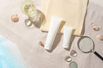 Top view of two white cosmetic tubes, seashells, a magnifying glass, a towel and glass balls displayed on a sandy background. Cosmetic sample for advertising.