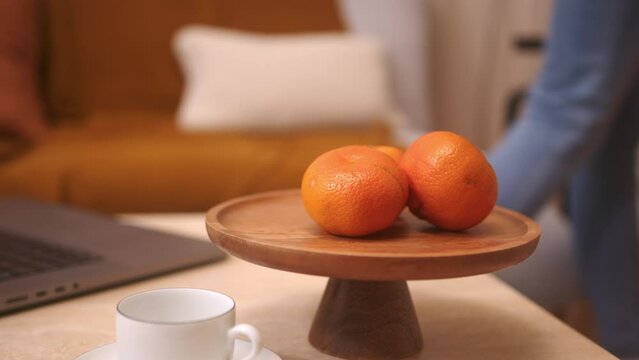 Close up shot of a table centerpiece with some mandarins on. On the living room table there is also a lalptop and furnishing objects. On the background there is a girl walks by. Slow mo