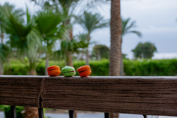 Macarons dessert in green and orange colors on the terrace, palms on the background. Sweet resort
