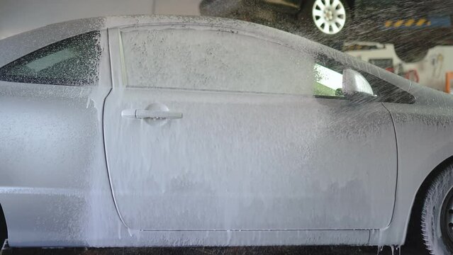 Auto detailer blankets car in luxurious foam, creating a captivating scene. The vehicle, immersed in suds, showcasing thorough cleansing artistry.