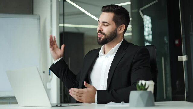 Successful bearded entrepreneur waving hello during online meeting on wireless laptop. Caucasian handsome man dressed in black suit sitting at office desk and enjoying distant communication.