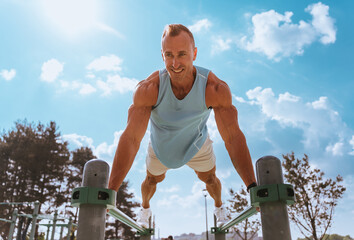 Focused and fit man in a sleeveless blue top executing perfect parallel bar dips under a clear blue...