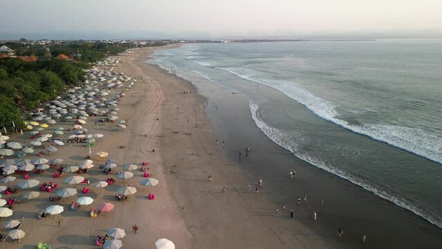 Aerial view of Beautiful sunset on the double six beach with Hotel, resort, cafe buildings, beach umbrella and calm ocean wave in Bali. Tourists are surfing, swimming and walking along the beach.