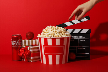 Popcorn in paper cup, gift boxes, soda, clapperboard and hand on red background