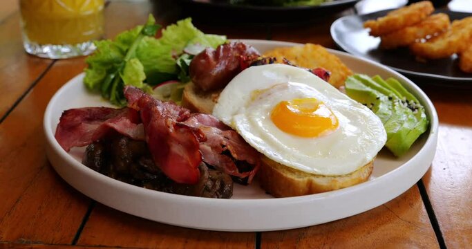 Big breakfast plate, close up view with selective focus, camera moving forward. Fried eggs on slice of bread, fried bacon and grilled sausage, sliced avocado and lettuce leaf, hash browns on back