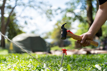 Man setting up camping tent outdoors, closeup. Human hand hammering the tent peg to the ground at the campsite.