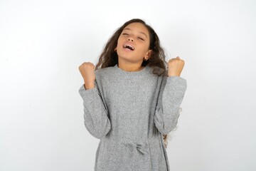 Beautiful teen girl wearing  grey dress over white background being excited for success with raised arms and closed eyes celebrating victory. Winner concept.