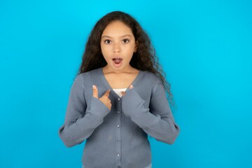 Embarrassed Beautiful teen girl wearing blue jacket over blue wall indicates at herself with...