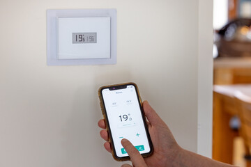 Person holding a smartphone in their hands to adjust their connected thermostat in a French house - 695295913