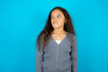 Obraz premium Amazed Beautiful teen gil wearing casaul clothes over blue background bitting lip and looking tricky to empty space.