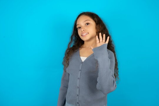Beautiful teen gil wearing casaul clothes over blue background Waiving saying hello happy and smiling, friendly welcome gesture.