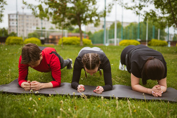 Group of fellows performing intensive plank exercise in gloomy green park. Friends try to stand in...