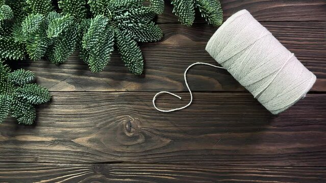 woman's hands putting twisted cotton rope, jute rope and scissors on dark wooden table decorated with spruce branches. Concept of handmade crafting, Christmas and winter leisure