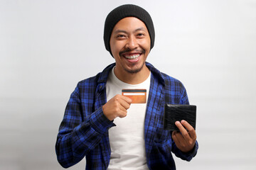 Excited young Asian man, dressed in a beanie hat and casual shirt, holds a wallet and credit card,...