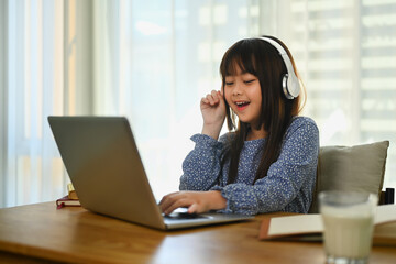 Smiling Asian little cute girl in headphone watching online lesson on laptop at home.