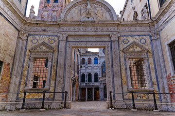 White stone gate with ornaments and weathered medieval stone facades of Scuola Grande di San Giovanni Evangelista at City of Venice on a cloudy summer day. Photo taken August 6th, 2023, Venice, Italy.