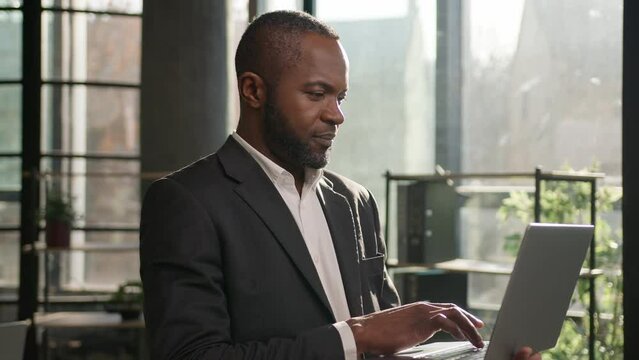 African American businessman ethnic adult 40s middle-aged business man stand with laptop in office sunny sunlight indoors. Smiling male employer CEO working computer online data analysis programming