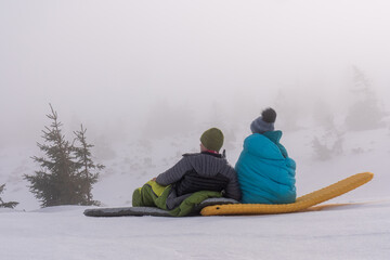A middle-aged woman and a man kiss and sit in sleeping bags on mattresses in the snowy mountains....