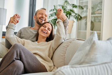 cheerful child-free couple holding hands and laughing with closed eyes on couch in living room, fun - obrazy, fototapety, plakaty