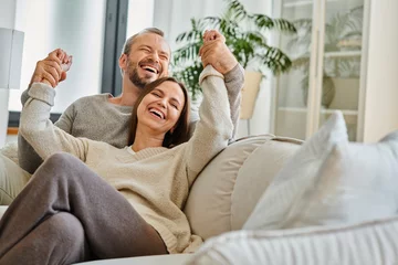 Fotobehang cheerful child-free couple holding hands and laughing with closed eyes on couch in living room, fun © LIGHTFIELD STUDIOS