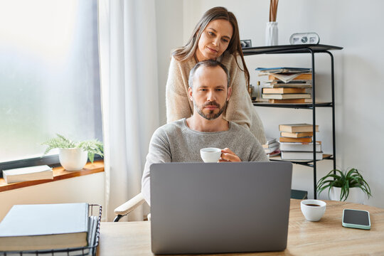 caring woman supporting thoughtful husband sitting with coffee cup at laptop in home office