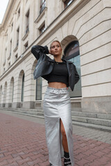 Beautiful young woman model in fashion outfit with a leather jacket with a skirt walks near a vintage building in the city