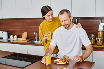Obraz na płótnie Canvas caring wife holding orange juice near pleased husband enjoying breakfast in kitchen, love and care