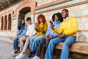 Group of happy diverse youth engaged on phones, sharing content in urban setting.