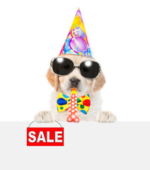 Funny Golden retriever puppy wearing sunglasses and party cap looks above empty white banner blows in party horn and shows signboard with labeled "sale"