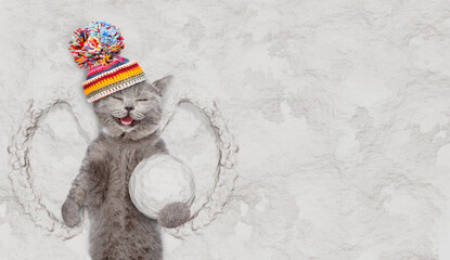 Happy cat wearing warm winter woolen hat with pompon holds big snowball while lying on snow. Empty space for text