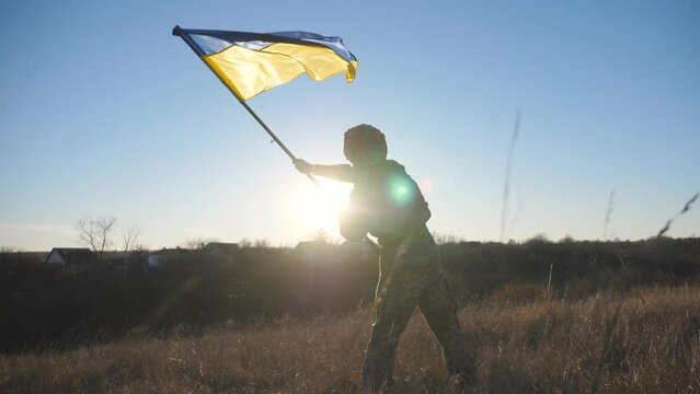 Dolly shot of male military in uniform waving flag of Ukraine at sunset. Young soldier of ukrainian army lifting national banner as symbol of victory against russian aggression. Invasion resistance