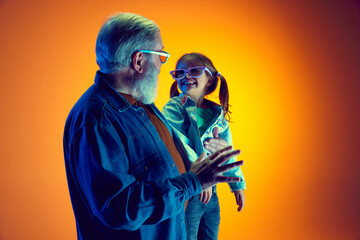 Senior man and little girl, grandfather and granddaughter watching TV over gradient orange...