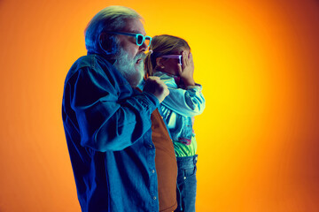 Horror movie. Senior man watching movie with little girl over gradient orange background in neon...