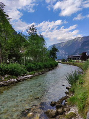 Fototapeta na wymiar A view on the Waldbach river, Austria. Stones on the shore. Mountain river stream. Green grass, coastline, trees. Blue sky, summer day in the Alps. Alpine village.