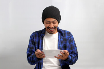 An excited young Asian man, dressed in a beanie hat and casual shirt, cheerfully holds an empty...