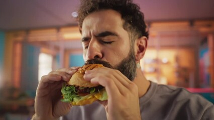 Fast Food Brand Commercial Concept: Slow Motion Portrait of a Young Stylish Man with a Beard Taking a Bite from a Delicious Hamburger, Looking at Camera and Smiling. Pure Enjoyment of Tasty Food