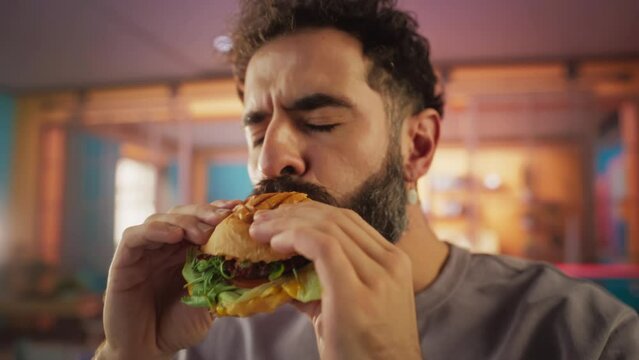 Fast Food Brand Commercial Concept: Slow Motion Portrait of a Young Stylish Man with a Beard Taking a Bite from a Delicious Hamburger, Looking at Camera and Smiling. Pure Enjoyment of Tasty Food