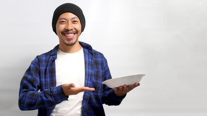 Excited young Asian man, dressed in a beanie hat and casual clothes, displays an empty white plate in the palm of his hand while standing against a white background