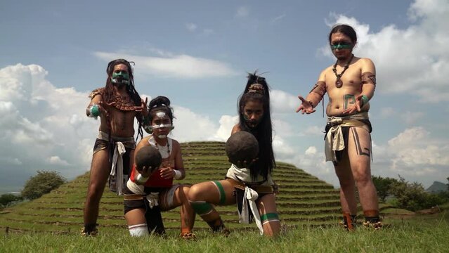 Group of serious Mayan warriors in traditional costumes with face painting holding black earthen balls during Mesoamerican ballgame while standing against green pyramid and looking at camera
