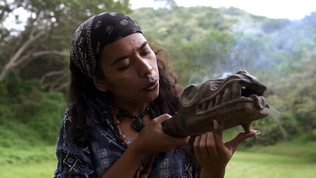 Focused young shaman woman in traditional Mayan attire performing a ritual smoking ceremony with a clay incense burner blowing copal smoke in Mexico