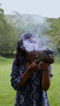Focused young shaman woman in traditional Mayan attire performing a ritual smoking ceremony with a clay incense burner blowing copal smoke in Mexico