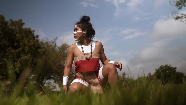 Young woman in ancient Mayan attire and white ceremonial pain in ger face crouched in a lush field in Mexico